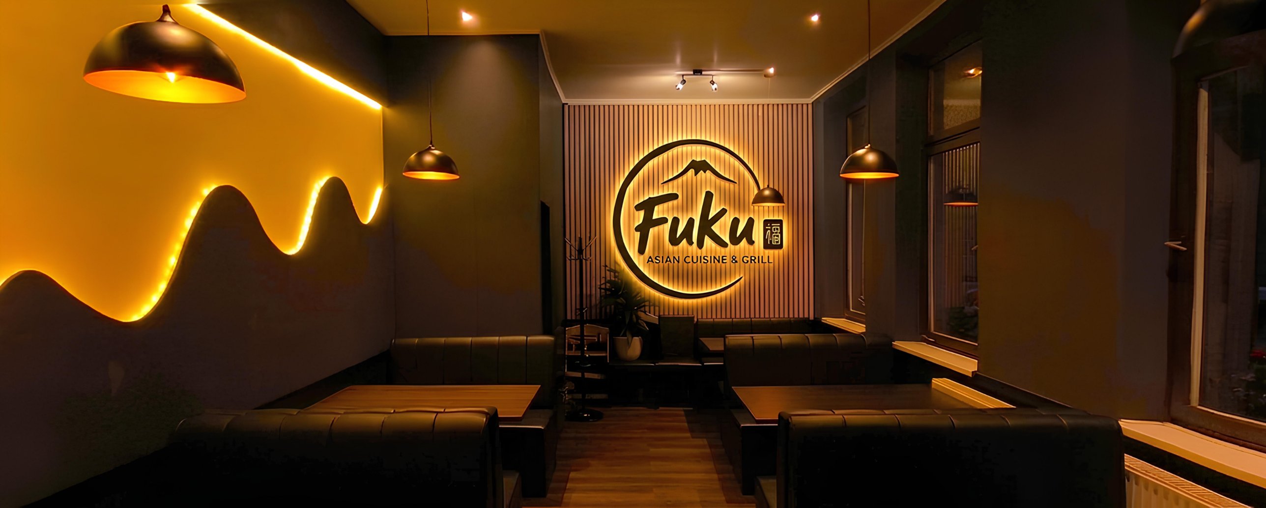 Gastraum des Fuku mit beleuchtetem Logo an der Holzlamellenwand, dunklen Sitzbänken und warmem Licht